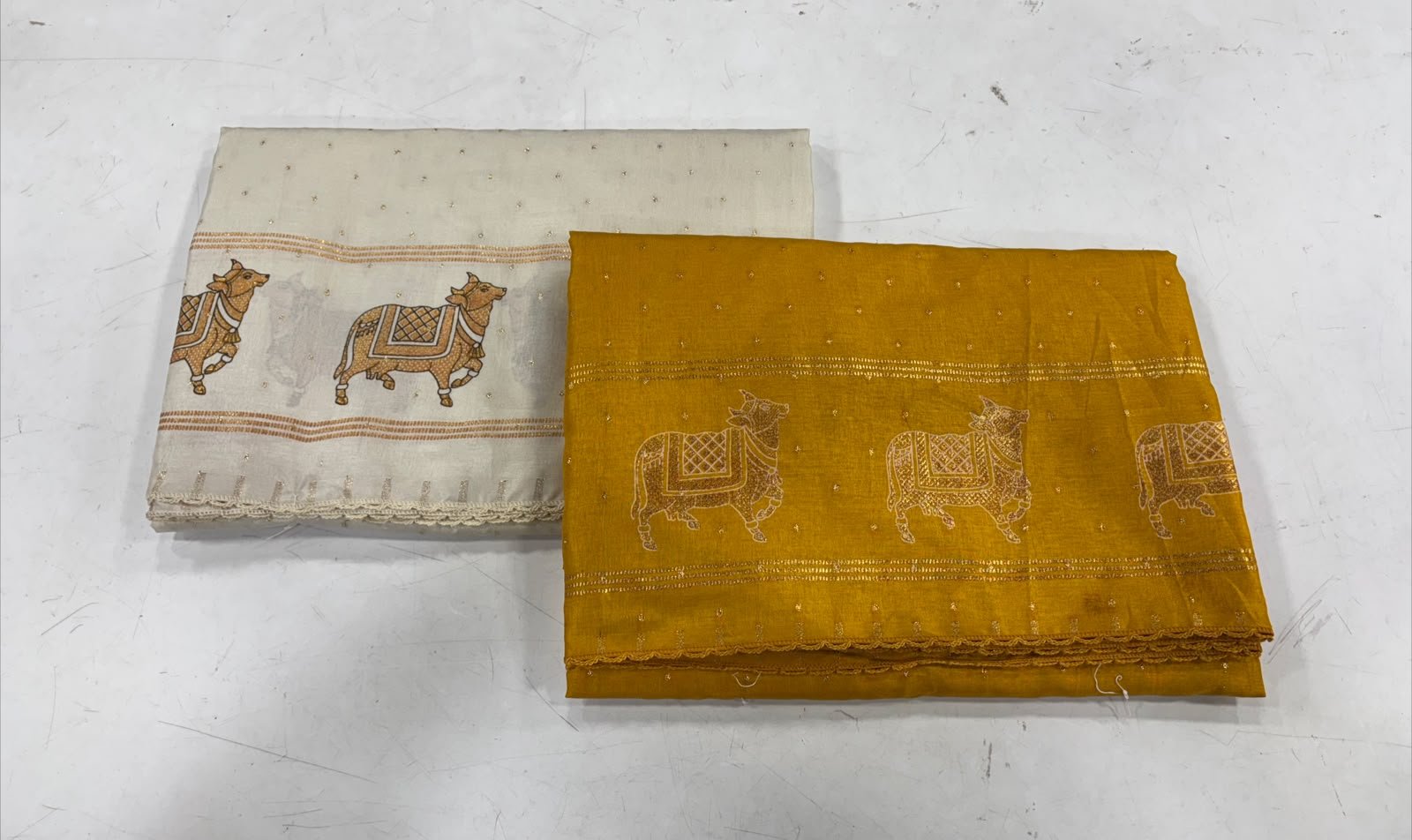 Two folded sarees featuring intricate gold cow motifs, presented in contrasting off-white and mustard yellow fabric.
