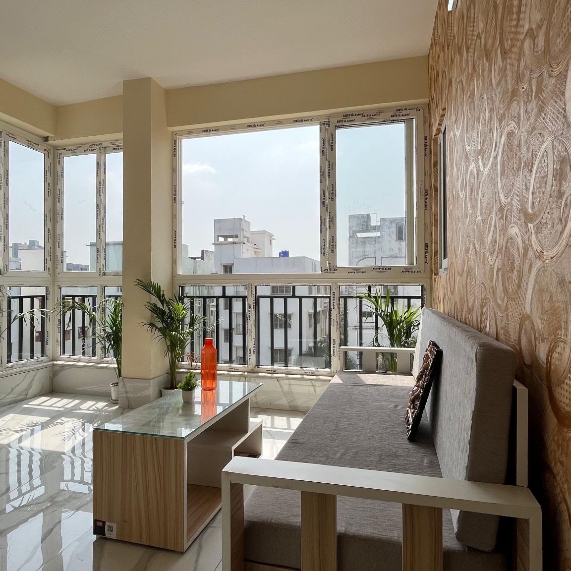 Sunlit lounge area featuring a grey sofa, glass-topped coffee table, and patterned wallpaper