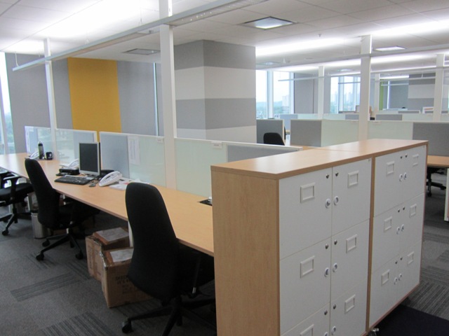 Modern open-plan office workspace featuring light wood desks, white storage lockers, and grey partition walls.
