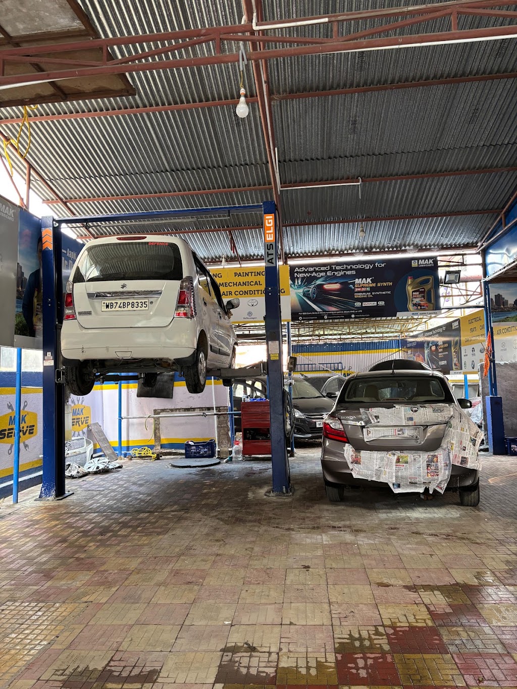 White Maruti Suzuki WagonR lifted on a hydraulic hoist inside the S.S Automobile workshop with a grey sedan undergoing bodywork in the foreground.