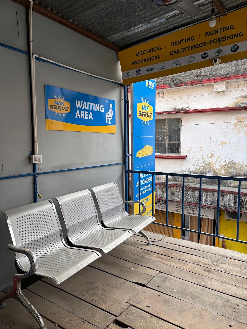 Waiting area at S.S Automobile featuring a row of three metal chairs beneath a blue and yellow signage board.